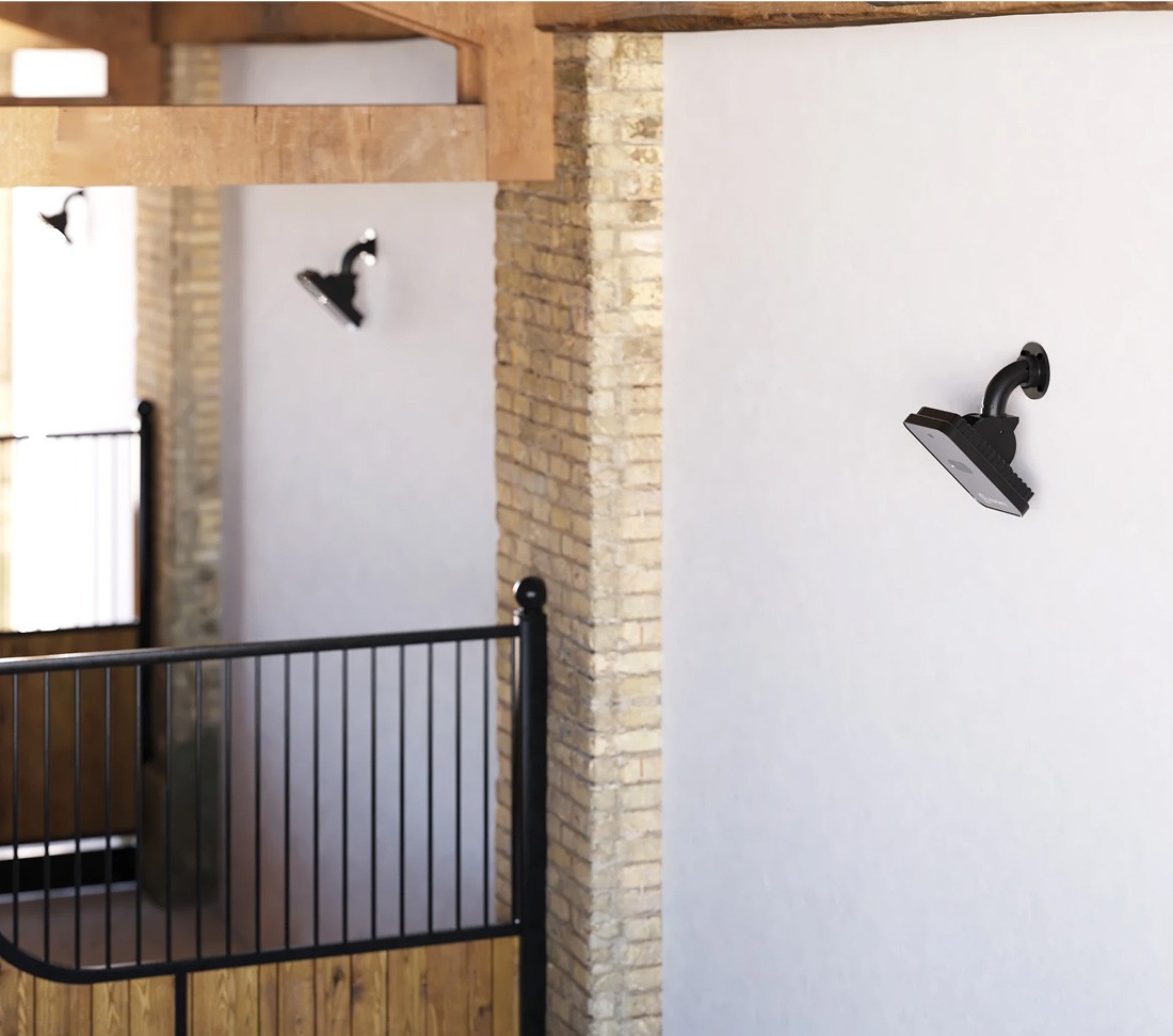 Close-up view of a premium stable interior with Horcery Stall Monitor devices mounted on white walls between brick pillars. The black monitoring units are angled downward toward individual stalls with wooden lower panels and black metal bars.